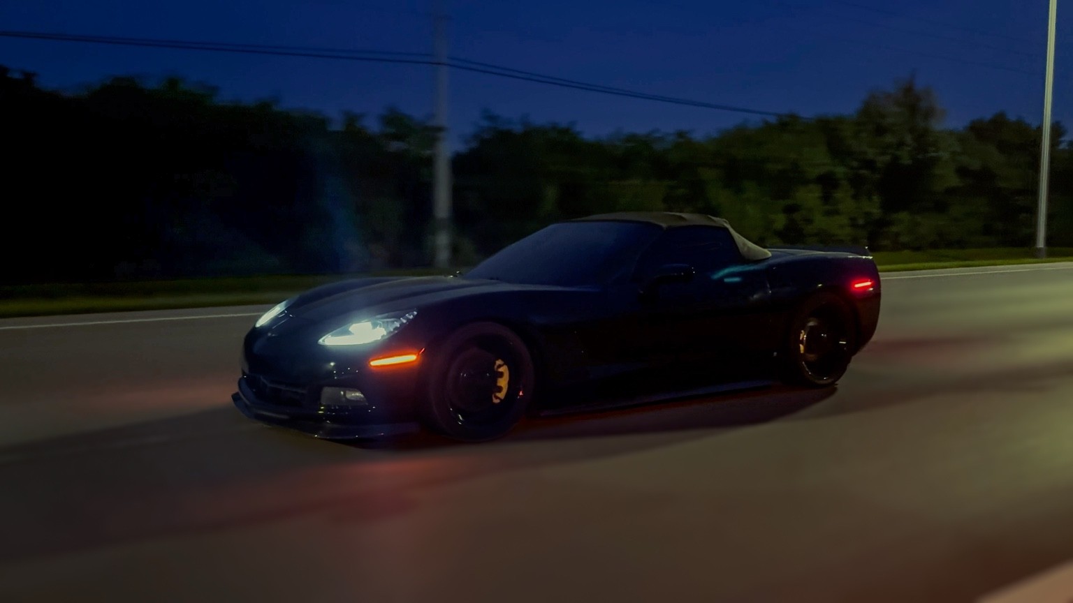 Black Corvette rolling along a road at dusk