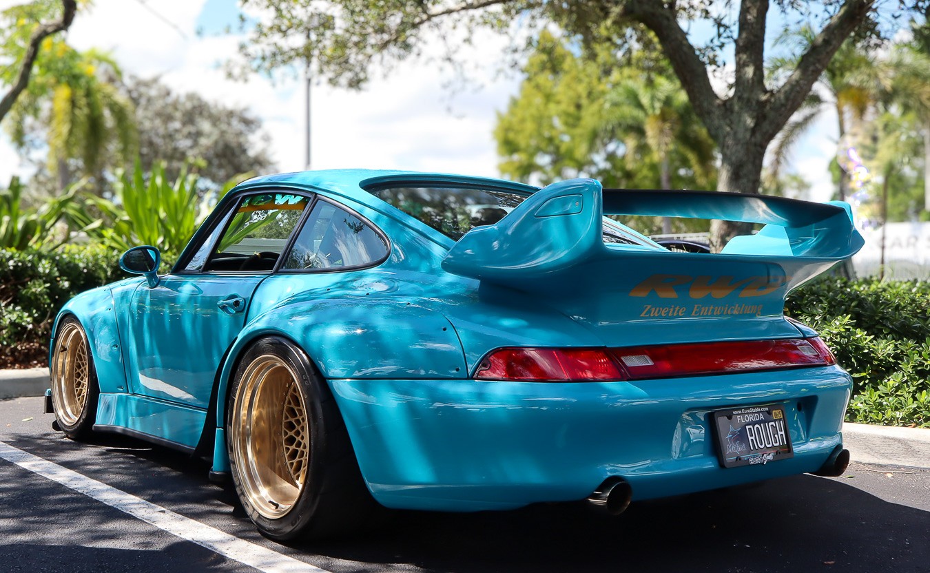 Turquoise RWB Porsche with widebody arches and a large rear wing beneath trees